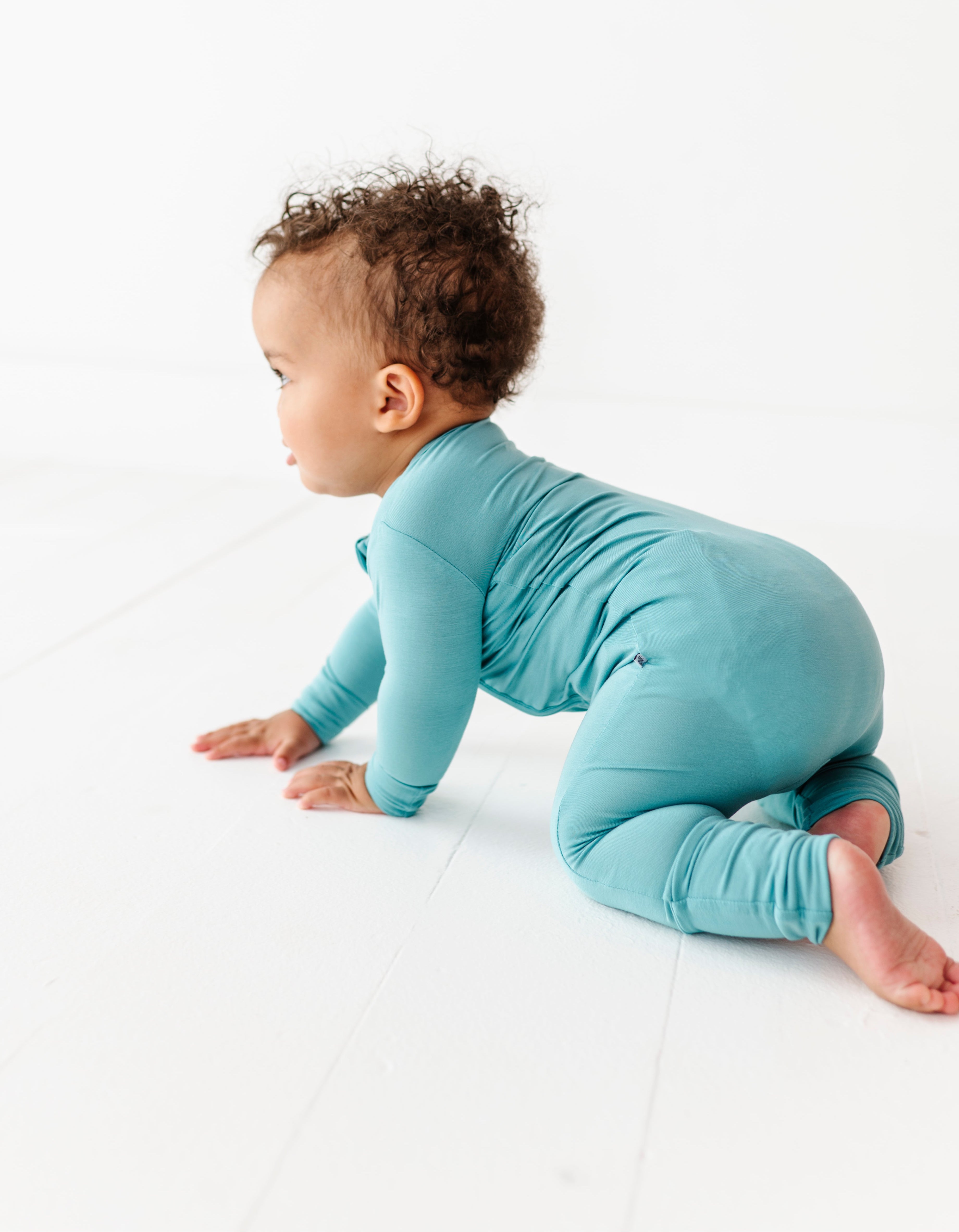 Baby in a slate blue convertible romper crawling on a white surface